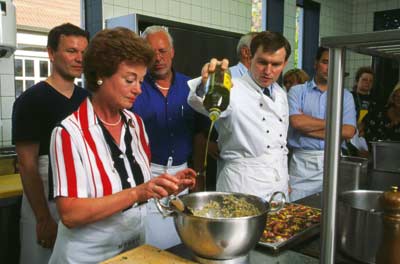 eine Gruppe besucht einen Kochkurs, der Kochlehrer gießt Olivenöl in die Teigmasse einer Dame 