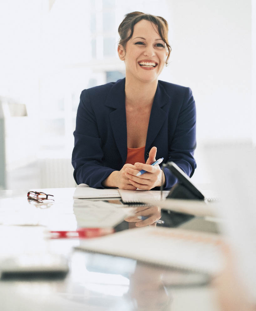 eine Geschäftsfrau sitzt im Blazer an einem Schreibtisch und lächelt 