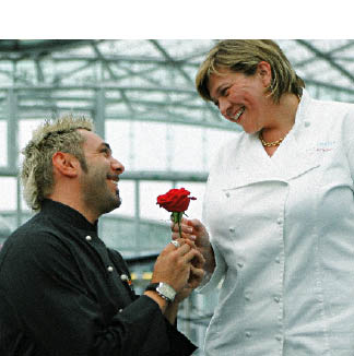 Ein Herr in einer schwarzen Kochjacke kniet vor einer Dame in weißer Kochjacke und schenkt ihr eine rote Rose 
