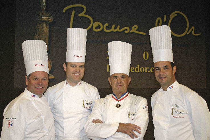 Paul Bocuse und Köche in Kochbekleidung posieren vor dem Logo des Bocuse d`Or