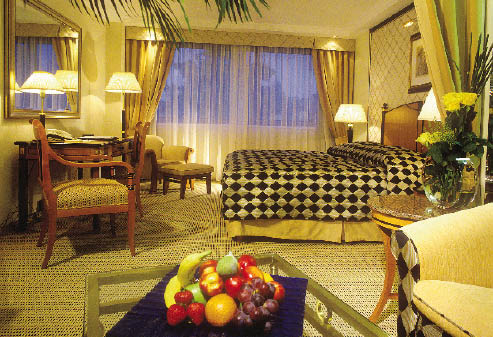 eine Suite im Kempinski, Ein Kingsize Bett vor einem Panorama Fenster, ein Schreibtisch in massiven Holz mit dem dazugehörigen Stühlen und ein Obstkorb auf dem Beistelltisch sind zu sehen 