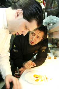 Roland Trettl mit einem Kollegen in der Küche, er trägt eine schwarze Uniform, der zweite Herr eine weiße beide lehnen über der Küchenzeile vor einem Teller 