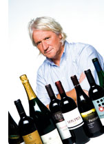 ein graumelierter Herr im blauen Hemd sitzt hinter einer Sammlung von Weinflaschen 