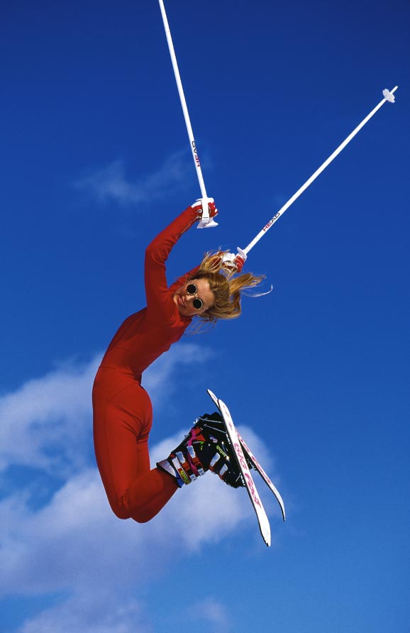 Eine Frau im roten Skianzug und Sonnenbrille springt mit Skiern und Skistöcken so hoch sie kann 