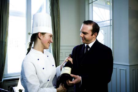 eine Dame in Kochgewand übergibt einem Herren in einem Anzug eine Champagnerflasche 