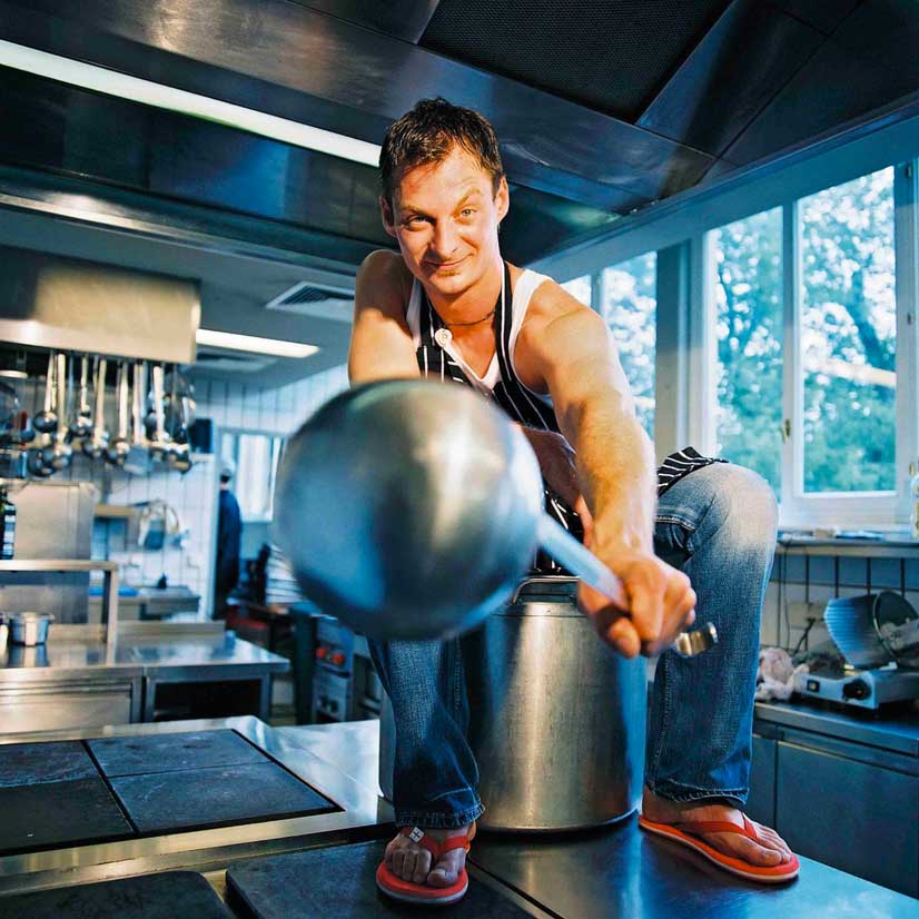 Bernie Rieder sitzt in der Küche auf einem Riesen Kochtopf mit einer Schöpfkelle in der Hand 