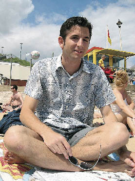 ein junger Herr sitzt in Shorts und Hemd am Strand 