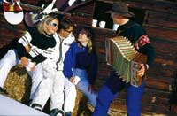 Ein Herr spielt Zierharmonika während drei junge leute fröhlich lauschen und vor einer Skihütte sitzen 