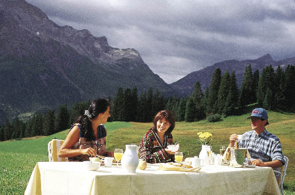 Ein Frühstückstisch auf einer Weide, im Hintergrund Berge und eine Familie die darauf frühstückt 