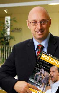 der Herausgeber des Rolling Pin Magazins steht im Anzug da mit einer Ausgabe in der Hand und lächelt in die Kamera