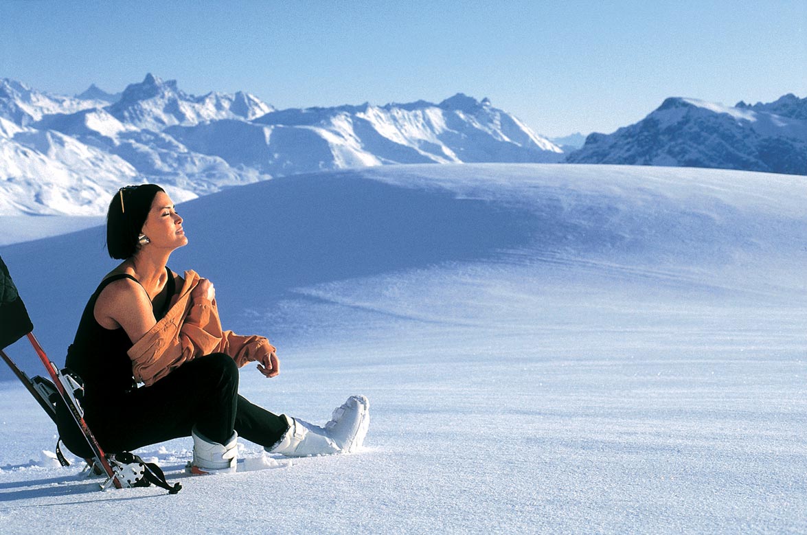 Eine Frau sitzt auf einem von Schnee bedecktem Berg und genießt die Sonnenstrahlen 