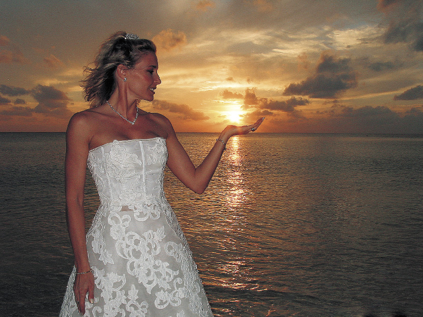 Eine Dame im Hochzeitskleid steht am Strand vor dem Sonnenuntergang und hält die Sonne in ihrer Handfläche 