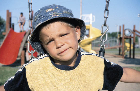 ein kleiner Junge sitzt auf der Schaukel am Kinderspielplatz und sieht fragend in die Kamera 